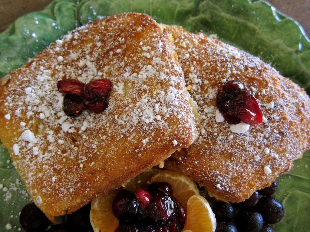 Two slices of Christmas French Toast sprinkled with powdered sugar, garnished with  macerated cranberries and served with tangerines, more cranberry and blueberries.